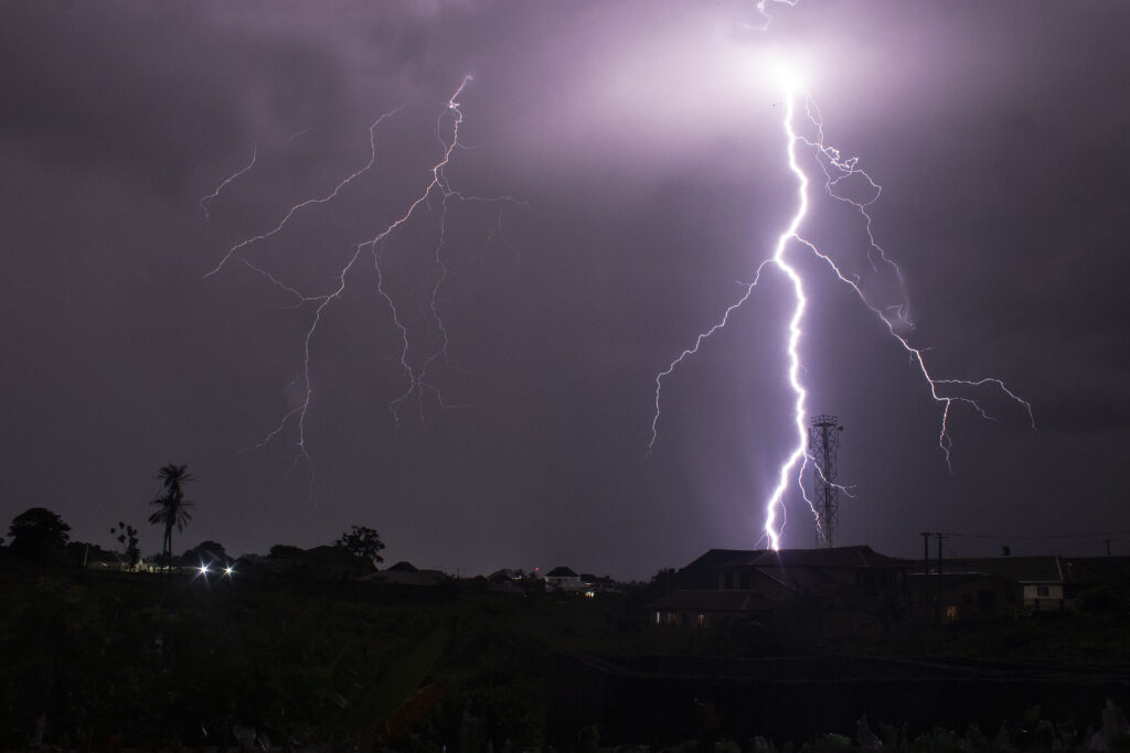 A powerful lightning bolt illuminates a dark, cloudy night sky over a rural landscape with palm trees and rooftops, creating an intense, dramatic scene.