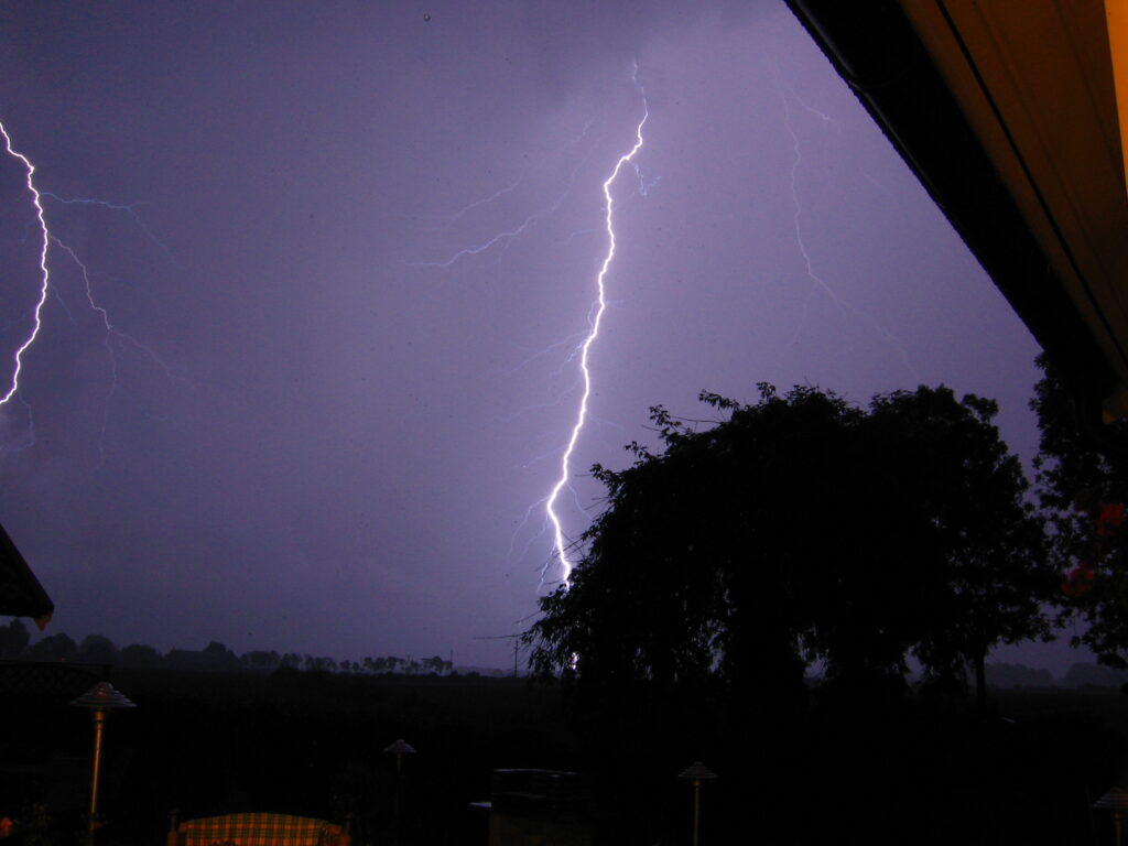 Dramatic night scene with a bright lightning bolt illuminating the sky, silhouetting trees and houses. The atmosphere feels intense and electrifying.