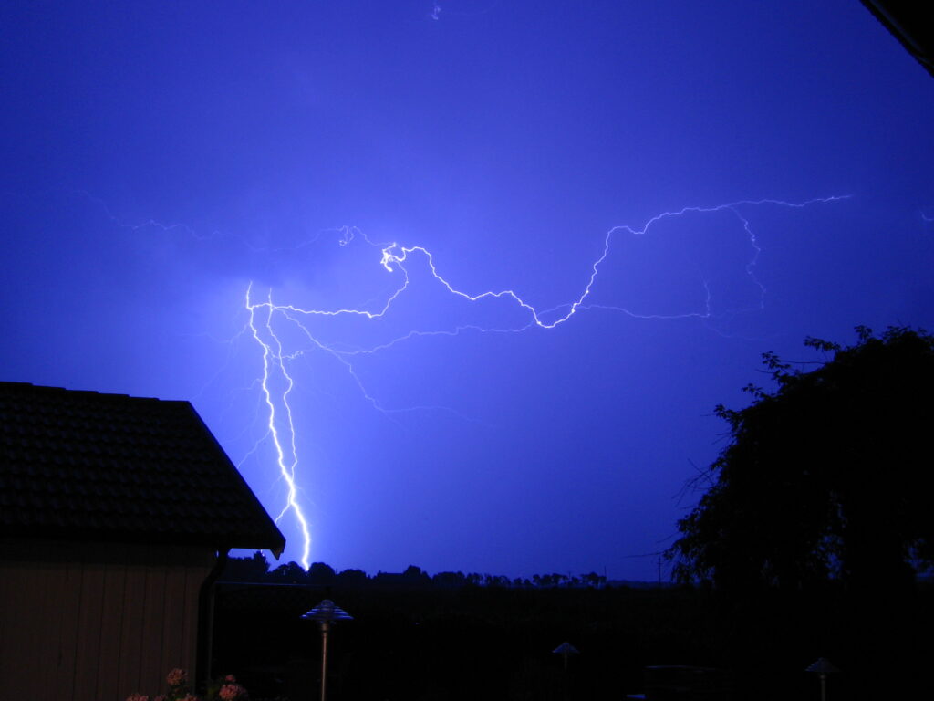 A vivid bolt of lightning illuminates a dark, blue night sky over silhouetted trees and a house with a tiled roof, creating a dramatic and electrifying scene.