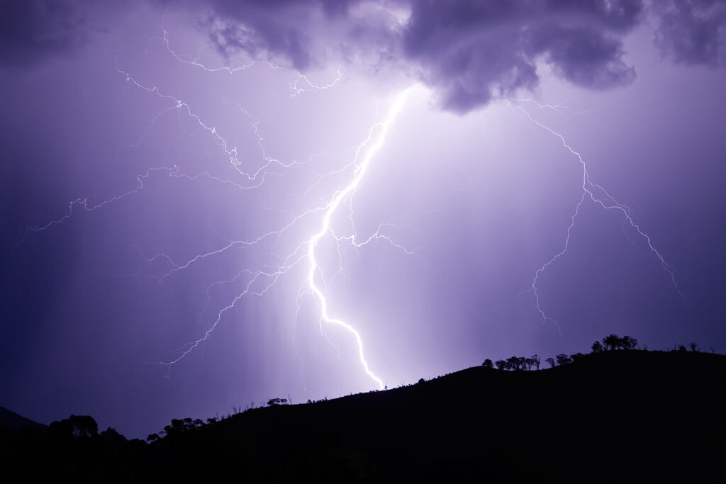 A bright lightning bolt illuminates a dark, cloudy sky above a silhouetted hilltop. The scene conveys intensity and the power of nature.