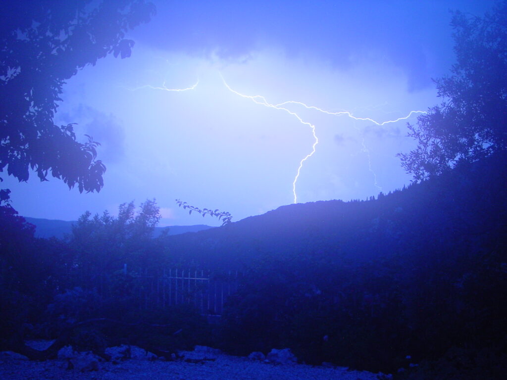 A vivid lightning bolt illuminates a dark, cloudy sky over silhouetted hills and trees, creating a dramatic and intense atmosphere in a nighttime landscape.