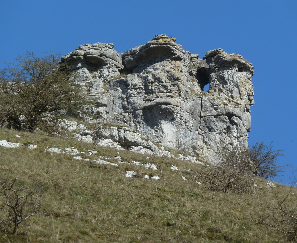Rock formation resembling a face with a hole for an eye stands under a clear blue sky. Sparse bushes and grass cover the slope below.