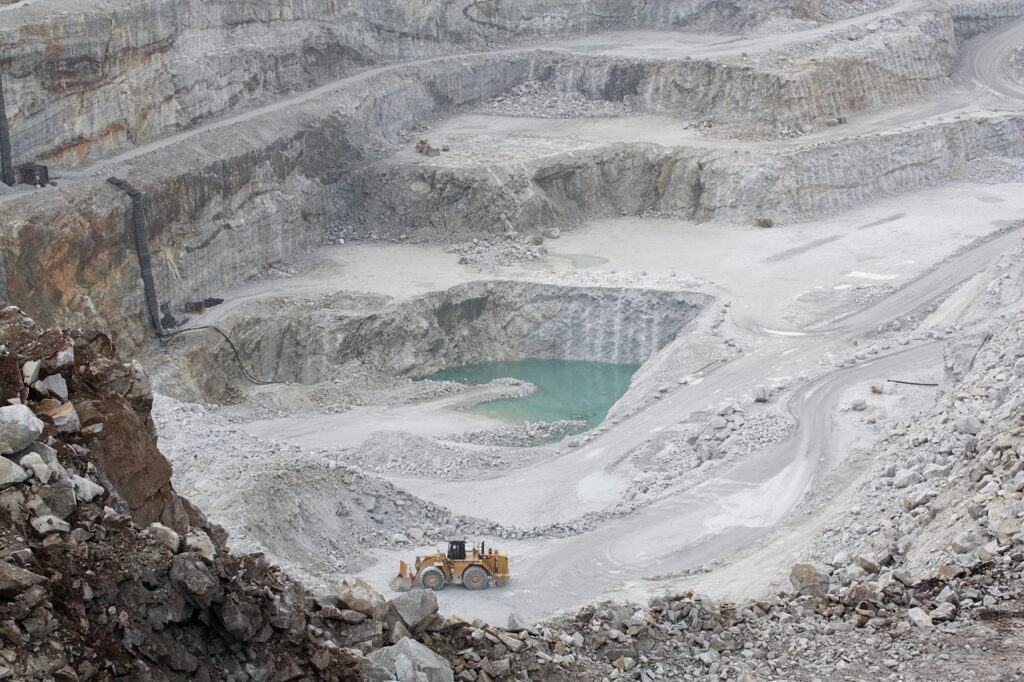 A vast quarry with layered rocky walls and a partially filled turquoise pool. A yellow bulldozer is visible, emphasizing the site's expansive scale.
