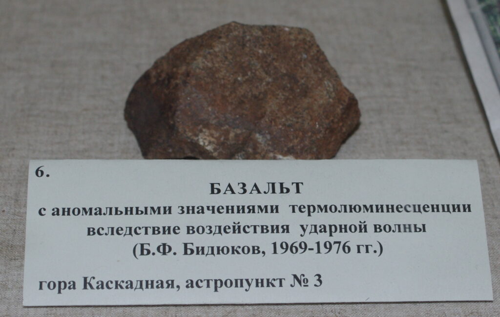 A rough, reddish-brown basalt rock is displayed on a beige surface. In front, a Russian label describes its anomalous thermoluminescence properties.