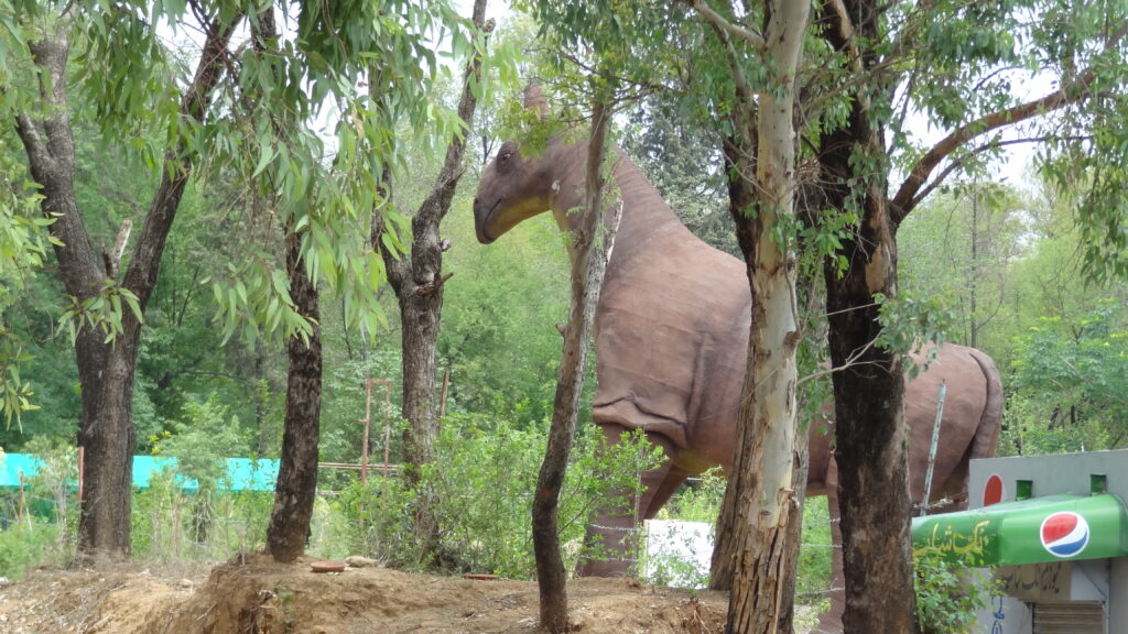 Large dinosaur sculpture surrounded by trees in a lush, green setting. The dinosaur is brown, with visible texture. Foreground features a Pepsi sign.