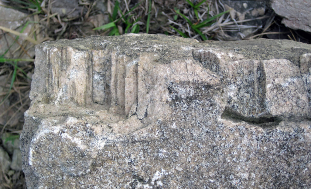 Close-up of a weathered stone fragment with vertical grooves resembling columns. Background shows grass and soil, suggesting an outdoor setting.