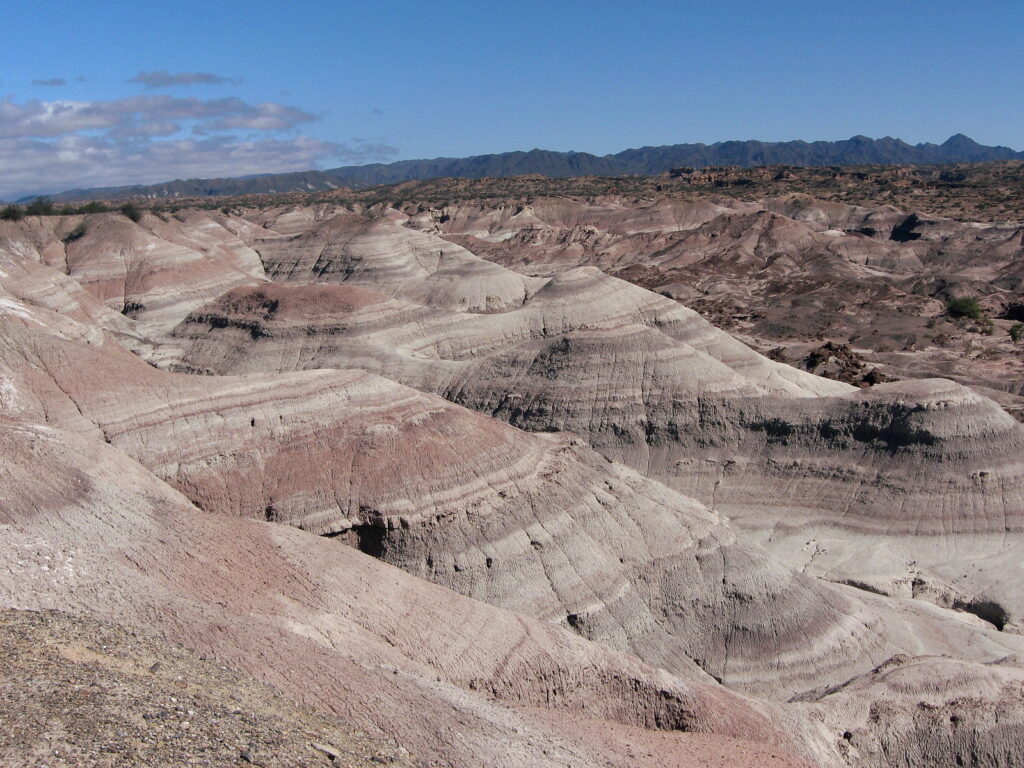 Sweeping view of pale, layered, eroded hills under a clear blue sky. Distant mountains line the horizon, evoking a serene, barren landscape.
