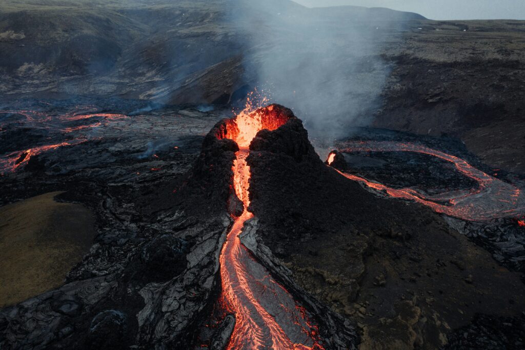 Aerial view of an active volcano spewing bright lava down its dark slopes, surrounded by barren, rocky terrain. Smoke rises, creating a dramatic scene.