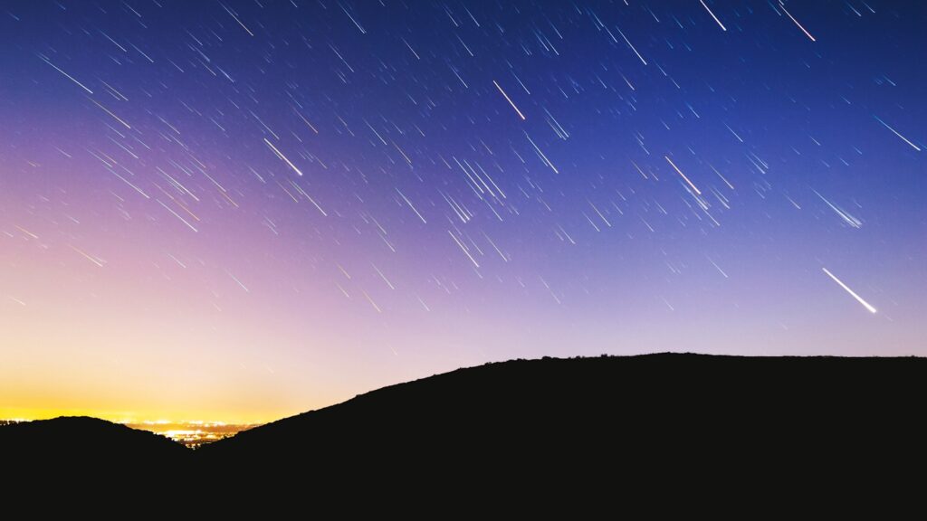 Starry sky with streaks of stars creating trails over a silhouetted mountain landscape. The horizon glows with city lights, casting a serene ambiance.
