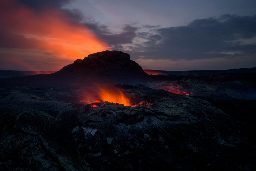 Molten lava glows brightly against a dark volcanic landscape at twilight. An orange-red sky adds drama and intensity to the scene.