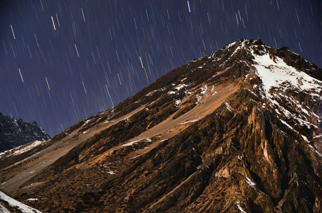 Mountain peak with snow patches under a starry night sky. Star trails arc across the scene, creating a serene and majestic atmosphere.