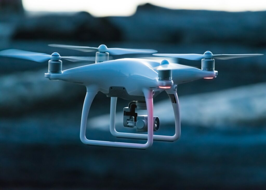 A white drone with four propellers hovers in a dark, blurred background. Red lights indicate its activity, creating a sense of motion and technology.