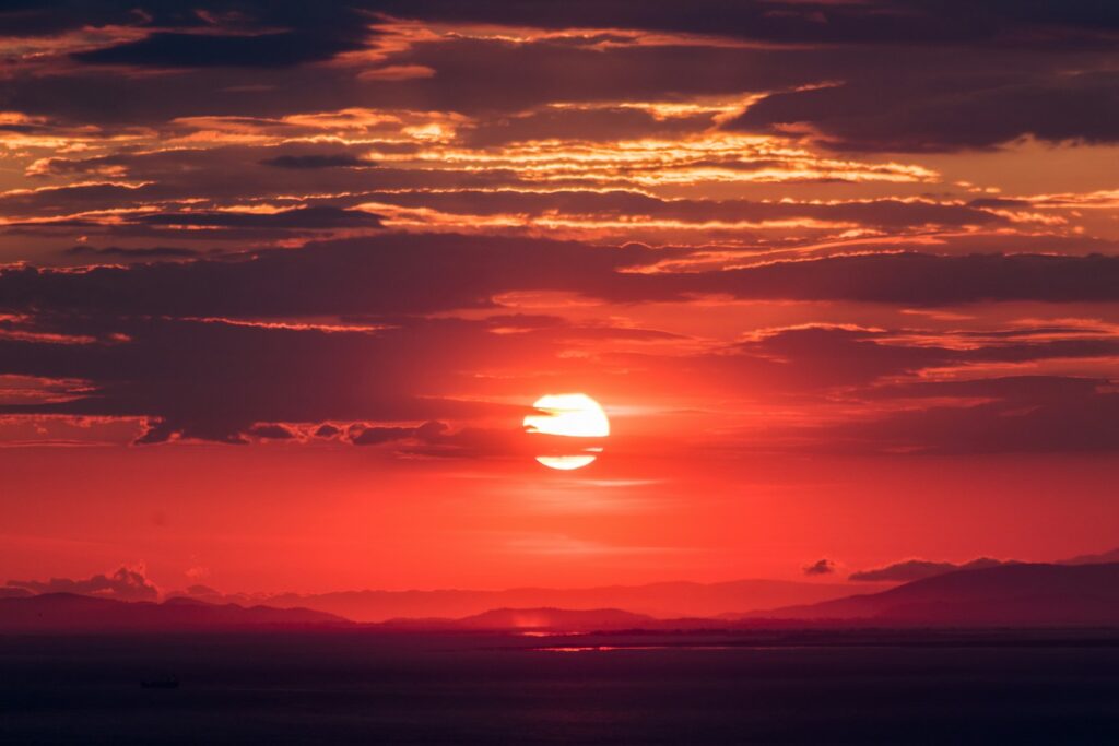 A vibrant sunset with a glowing red sun partly obscured by dark clouds fills the sky. The sea and silhouetted hills are visible beneath, evoking tranquility.