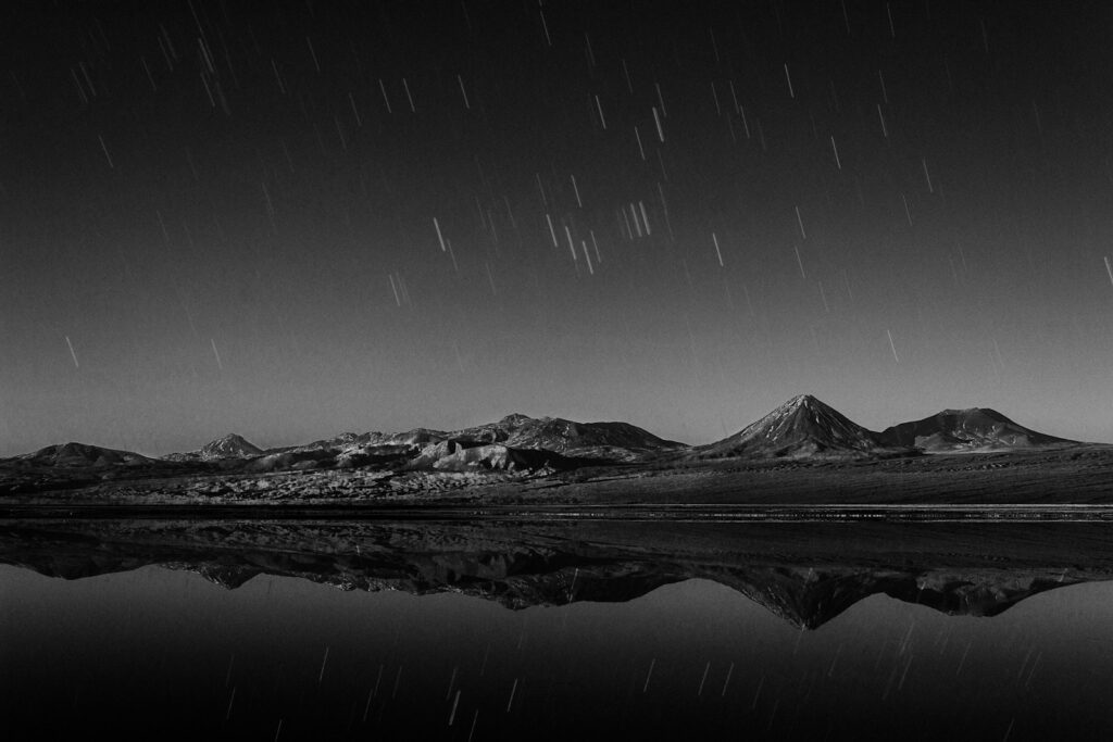 Black and white image of star trails over a calm lake reflecting distant mountains, creating a serene and timeless atmosphere.