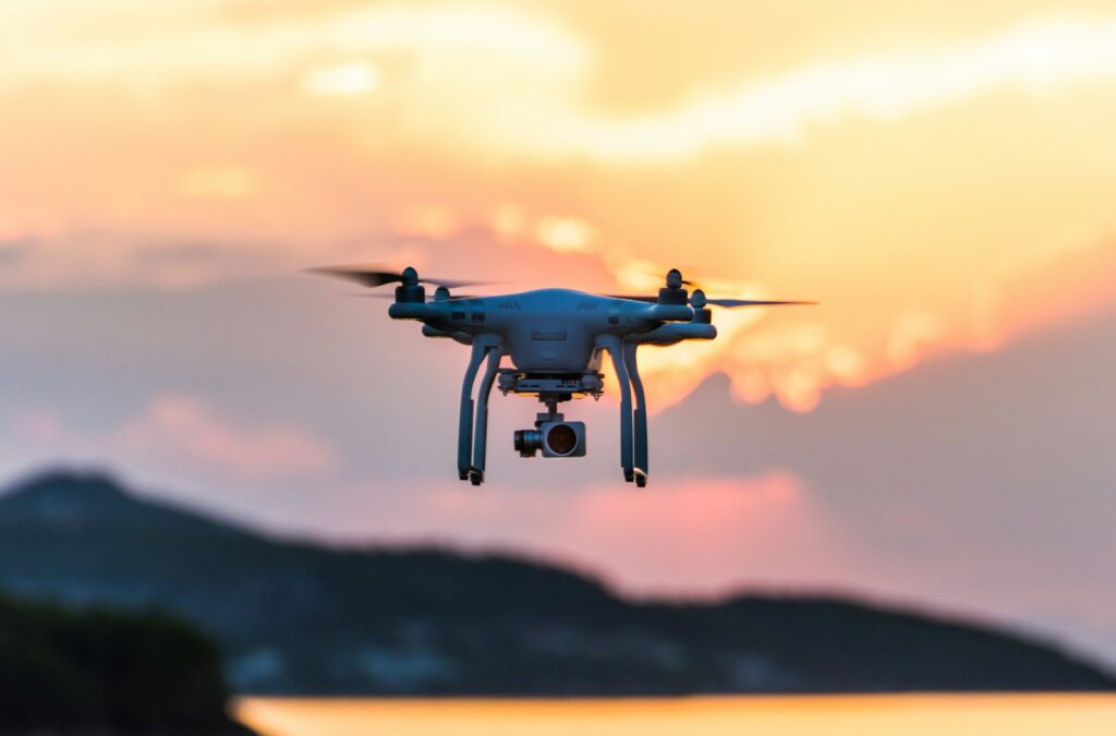 A drone hovers mid-air against a vibrant sunset, with orange and pink hues. Silhouetted mountains and calm water are visible in the background.