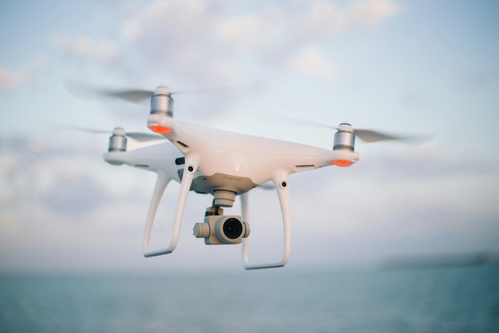 A white drone with four propellers hovers against a serene sky and ocean backdrop. The scene evokes a sense of calm and technological advancement.