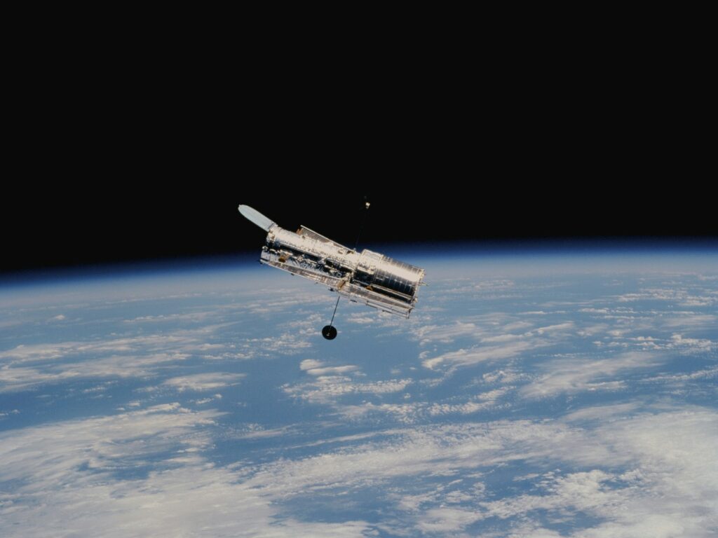A telescope orbits above the Earth, partially illuminated by sunlight. Below, a curve of the Earth's horizon with white clouds set against a deep, dark space.