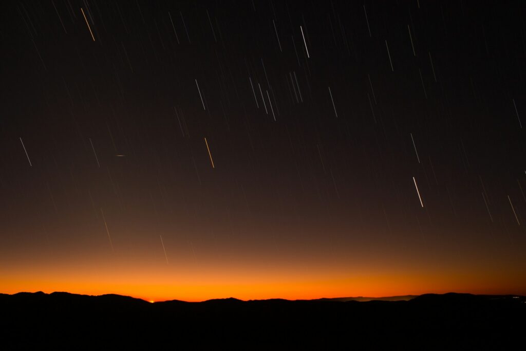 Starry night sky with long trails over a dark mountain range. The horizon glows warm orange, suggesting dusk or dawn, creating a serene atmosphere.
