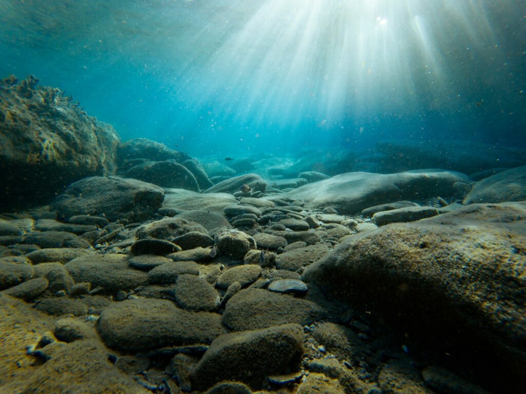 Underwater scene with sunlight streaming through clear blue water, illuminating smooth rocks on the seafloor. A calm, serene, and tranquil atmosphere.