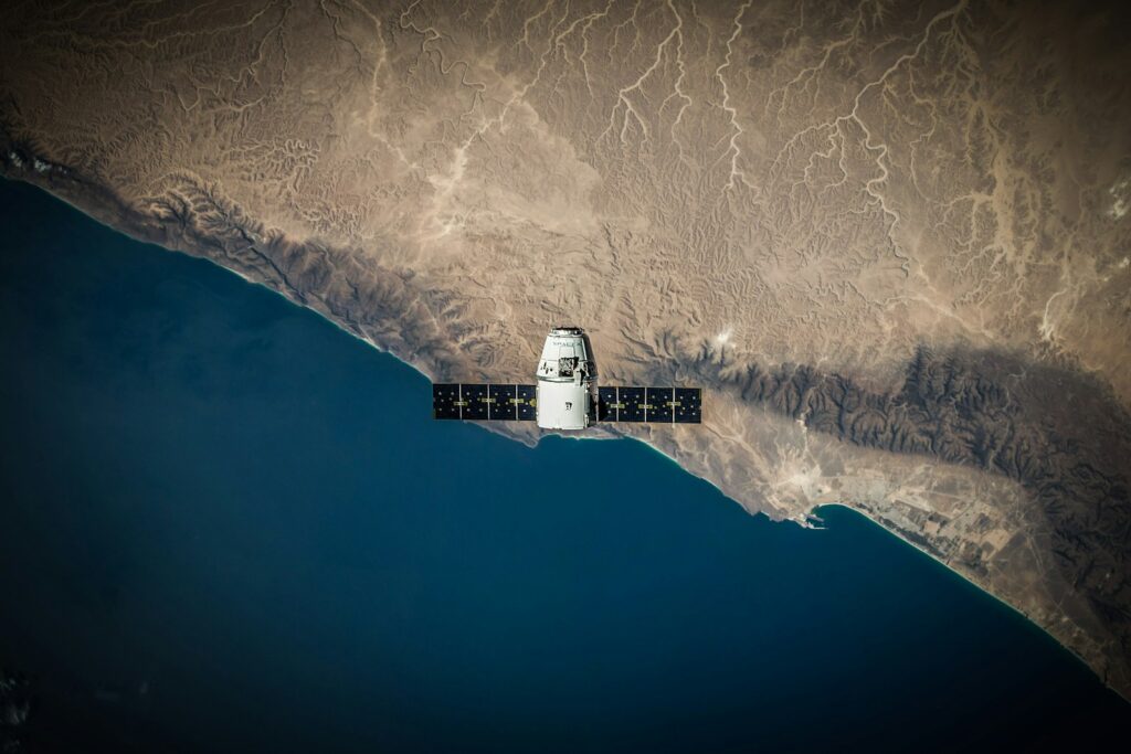 Satellite with solar panels orbits Earth, spanning a desert landscape against a deep blue ocean. The scene conveys a sense of exploration and technology.