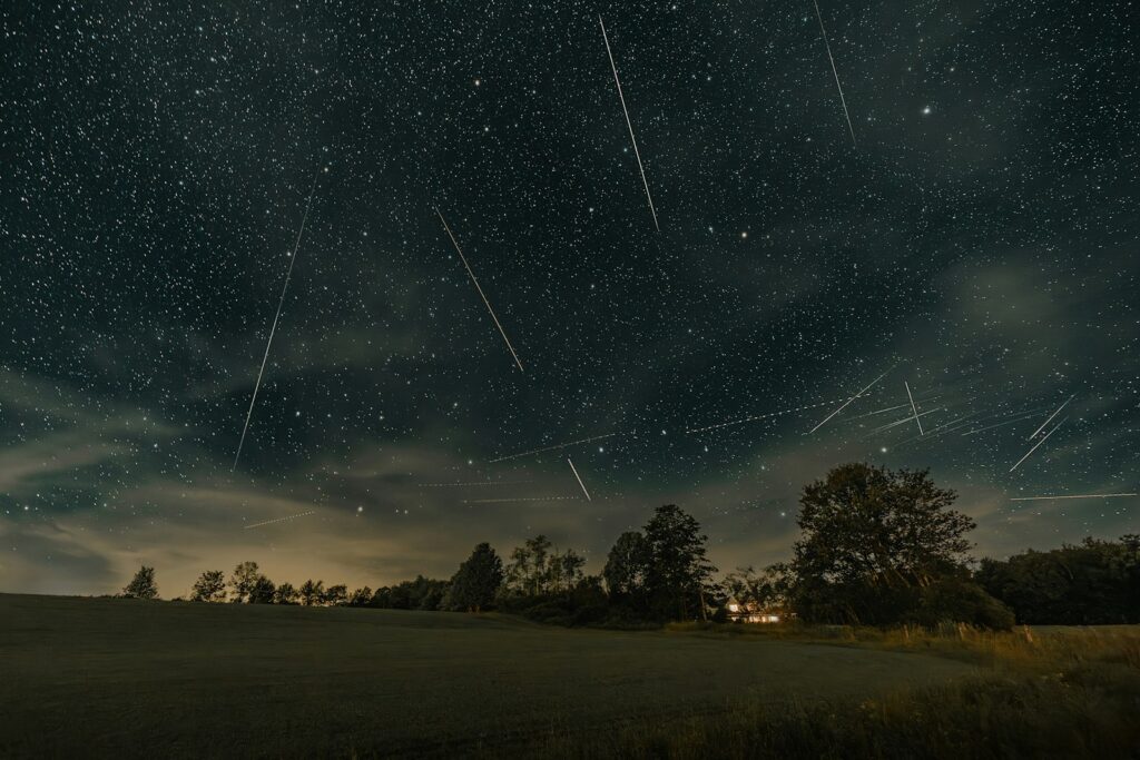 Night sky filled with numerous shooting stars above a peaceful grassy field and silhouetted trees. A small house emits warm light, conveying tranquility.