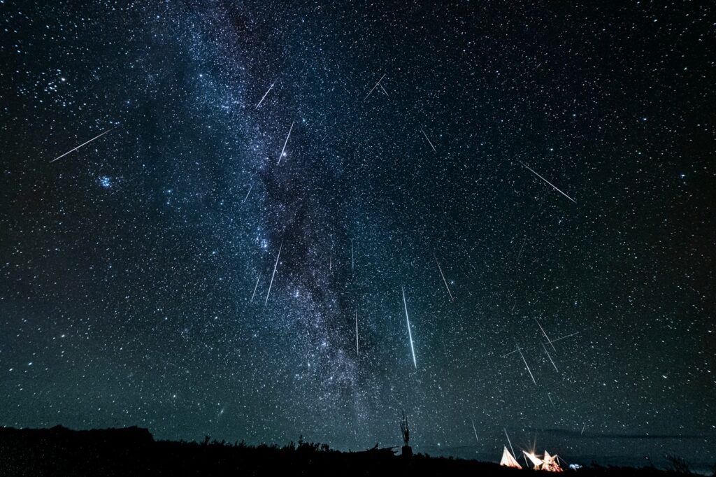 Star-filled night sky with bright meteors streaking across, set against the Milky Way. Silhouetted tents and trees create a peaceful camping scene.