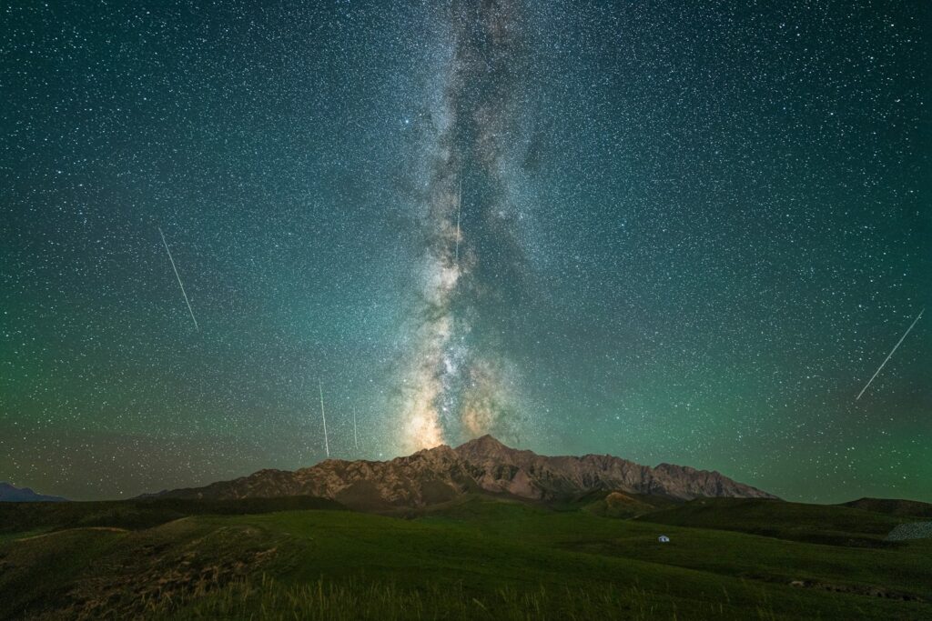 Starry night sky over mountains with the Milky Way clearly visible. Shooting stars streak across, creating a serene and awe-inspiring scene.