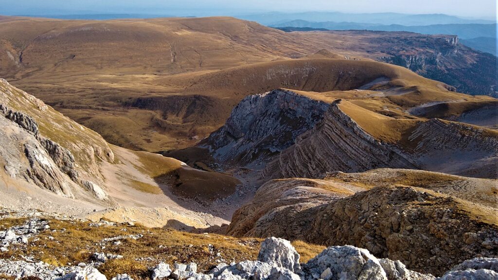 Rocky mountain landscape with rugged cliffs and grassy plateaus under a clear blue sky. The mood is serene and expansive, invoking a sense of wonder.