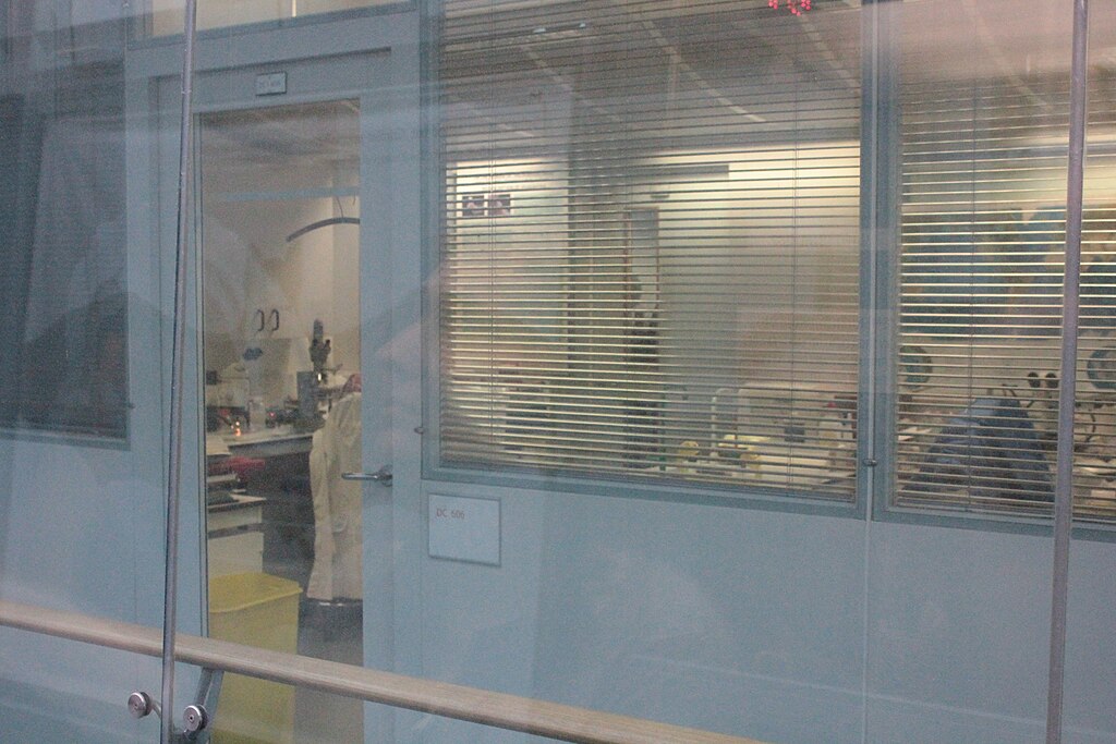 A laboratory room viewed through glass with blinds. Inside, a lab coat and scientific equipment like microscopes are visible. The scene is calm and sterile.