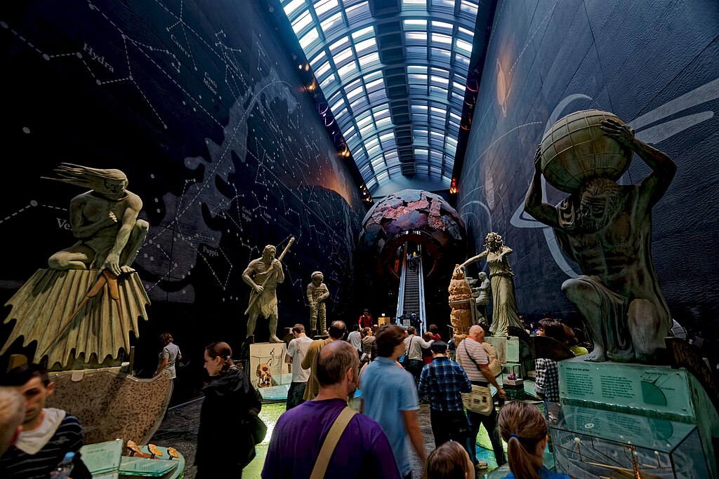 Visitors walk through a museum exhibit featuring large statues of mythological figures under a high-arched ceiling with a cosmic map on the walls.