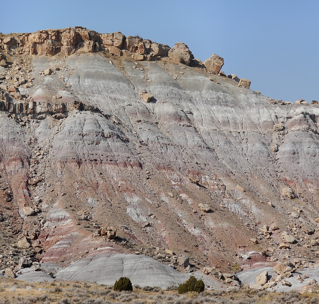 Rocky desert landscape with stratified hills in gray and red hues under a clear blue sky. Sparse shrubs dot the barren terrain, exuding a rugged and serene atmosphere.