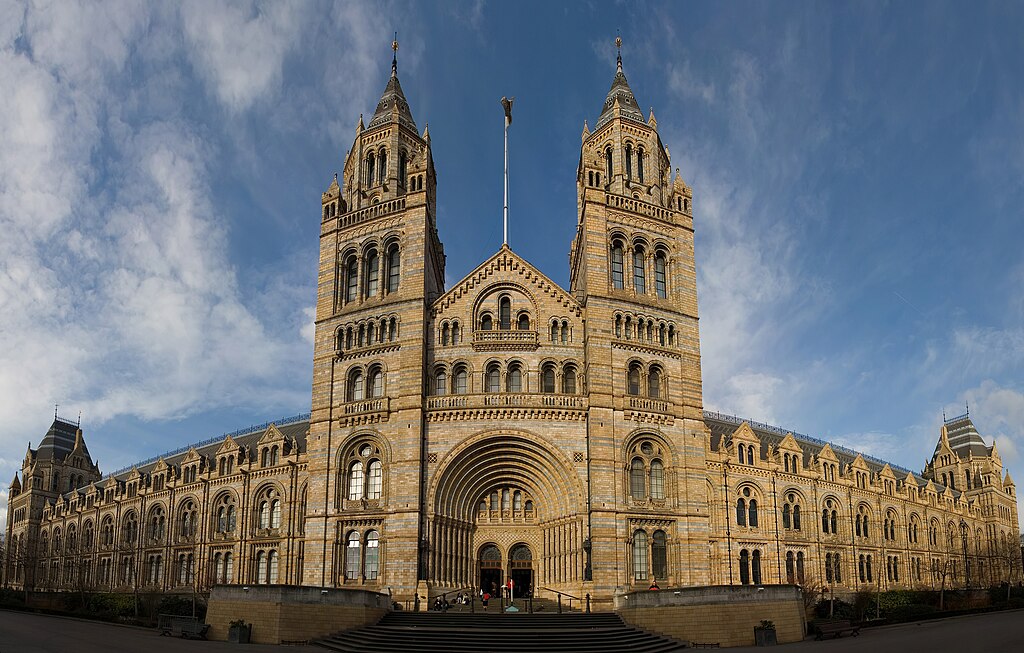 A grand historic building with ornate architecture, featuring two tall towers and intricate stonework, set under a vibrant blue sky with scattered clouds.