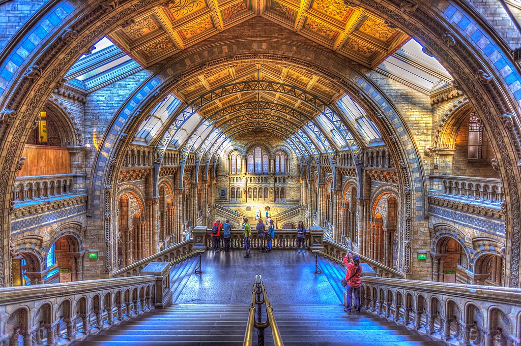 Grand hall of a historic building with ornate arched ceilings, stone columns, and large windows. Visitors admire the architecture, creating an awe-inspiring atmosphere.