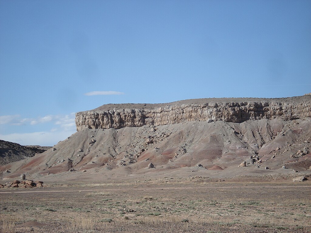 Rocky desert cliff under clear blue sky. The sunlit cliff exhibits horizontal rock layers and slopes down to a barren foreground, evoking solitude and vastness.
