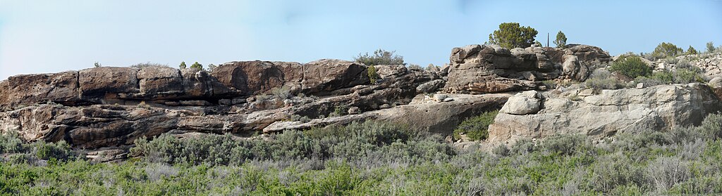 Panoramic view of rugged, weathered rock formations with sparse greenery in the foreground under a clear blue sky, evoking a serene desert landscape.