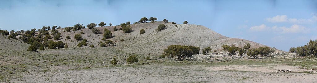 Arid landscape with sparse green vegetation on rolling hills under a clear blue sky. Scattered trees dot the dry terrain, creating a tranquil scene.