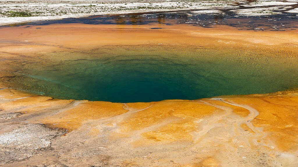 A vibrant hot spring with a deep blue center, surrounded by colorful orange and yellow mineral deposits, set in a rocky terrain under a clear sky.