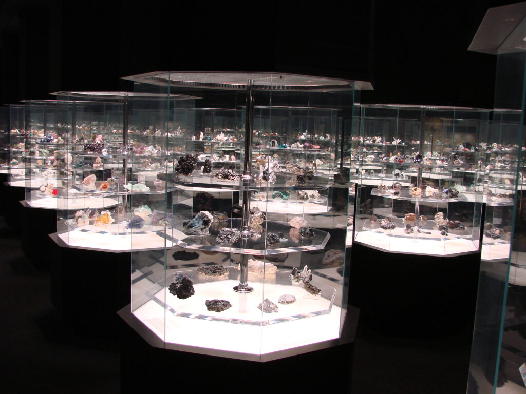 A dimly lit museum exhibit showcases numerous minerals and gems in sleek glass cases. The colorful stones are carefully arranged and illuminated.