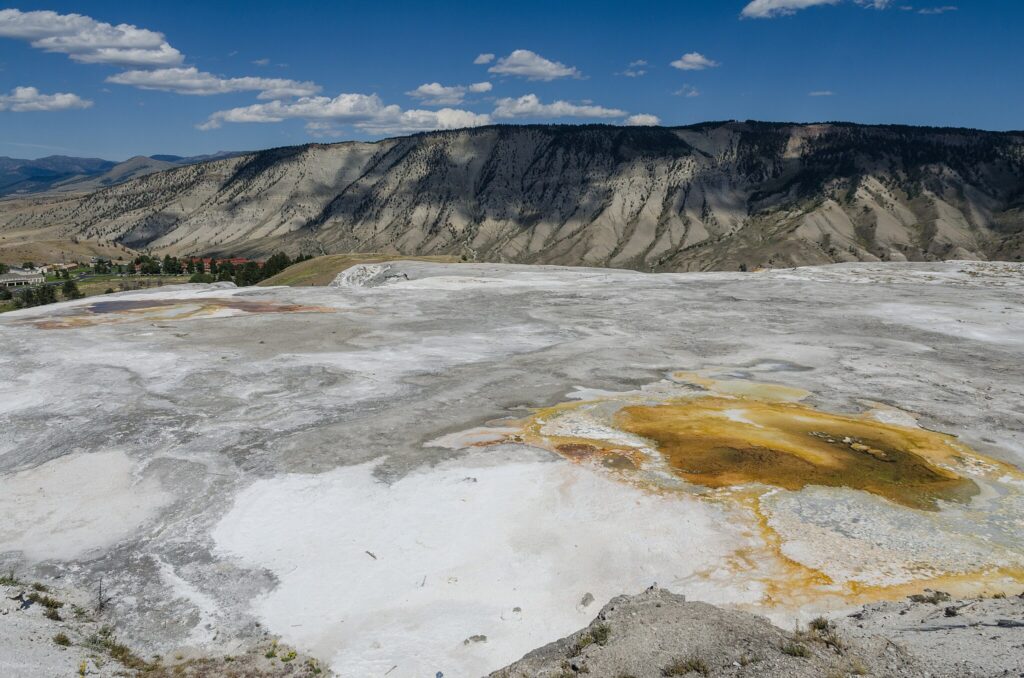 Expansive rocky geothermal landscape with white, gray, and yellow hues. Mountains stretch across the horizon under a clear blue sky with scattered clouds.