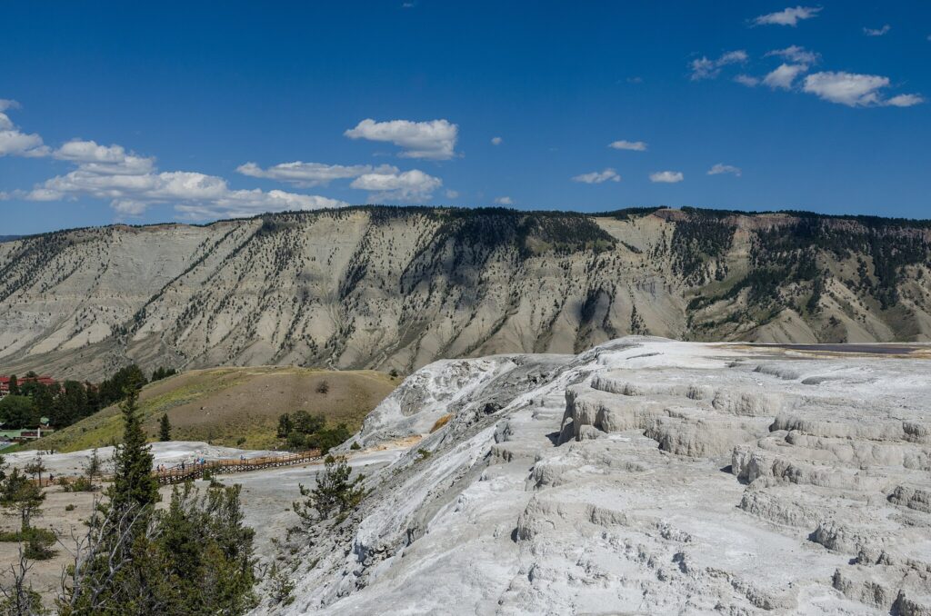 Rocky, white limestone terraces dominate the foreground, with a mountainous backdrop under a clear blue sky. A boardwalk is visible in the distance.