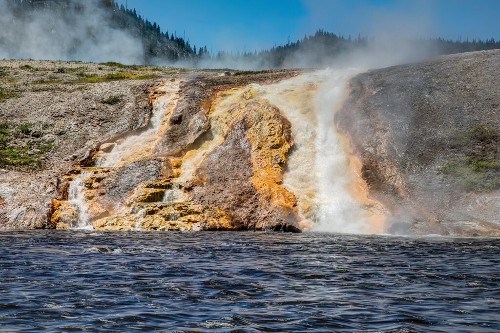 Steaming hot spring cascades down rocky terrain into dark rippling water, with rising steam and distant pine-covered hills under a clear blue sky.