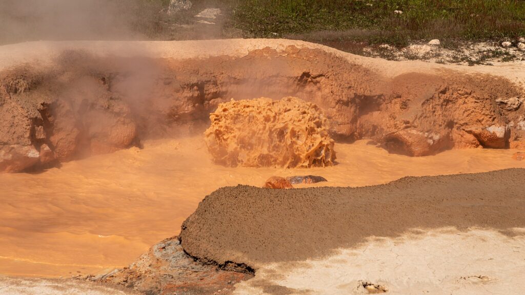 Bubbling mud pool with steam rising, surrounded by cracked, reddish clay. The scene conveys a sense of raw geothermal energy and natural heat.