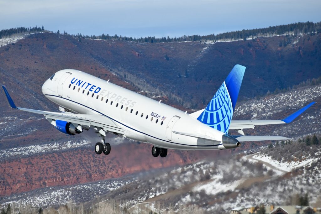 A Embraer E175 airplane takes off against a backdrop of snow-dusted mountains. The blue and white aircraft ascends into a clear sky.
