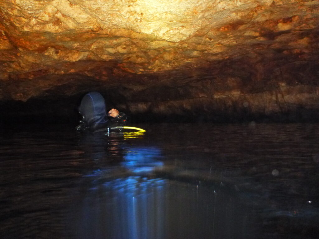 A diver in a wetsuit explores a dimly lit, orange-tinged underwater cave, creating a mysterious and adventurous atmosphere. Reflections shimmer on the water's surface.
