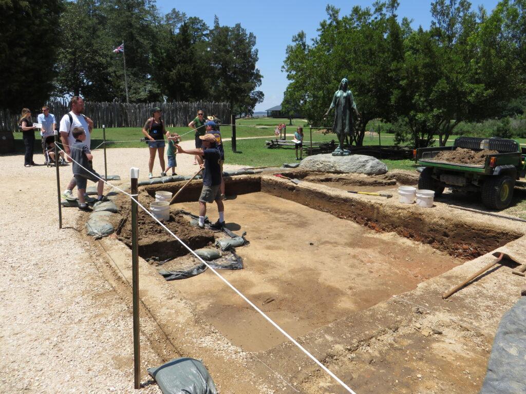 Kids and adults observe an archaeological dig in a historic park, with a statue in the background and a vehicle with tools nearby, under a sunny sky.