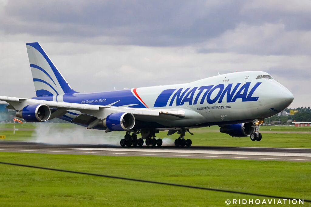 Boeing 747 landing on a runway with smoke from wheels. The aircraft is branded "National" with blue and white colors. Overcast sky and green grass surround.
