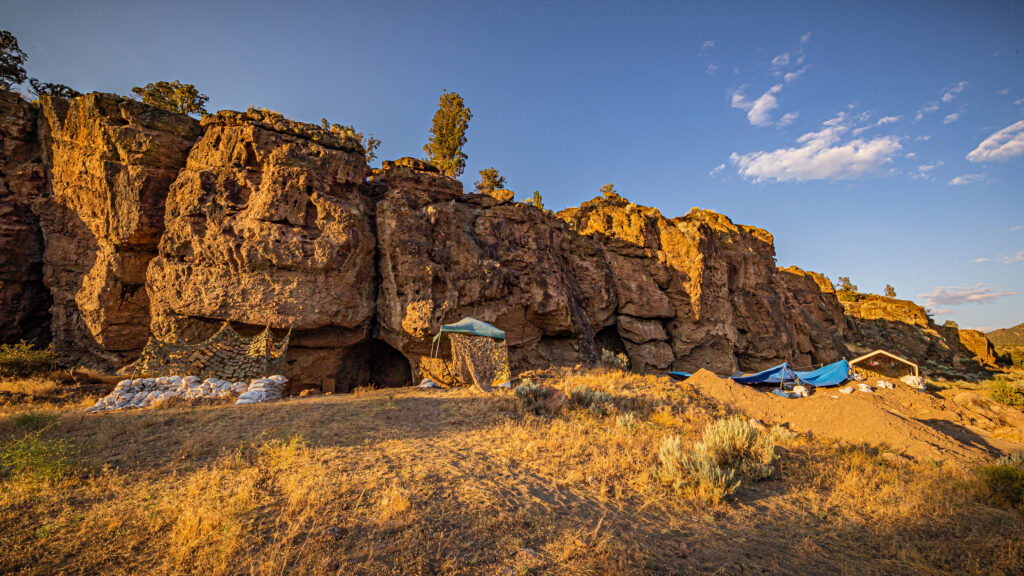 Rocky cliffs bathed in warm sunlight with a clear blue sky. Tents and tarps are nestled at the base, suggesting an archaeological dig site.