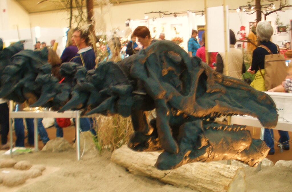 A large dinosaur skeleton replica is displayed on a sandy exhibit surrounded by people. The scene is vibrant, with an educational and curious atmosphere.