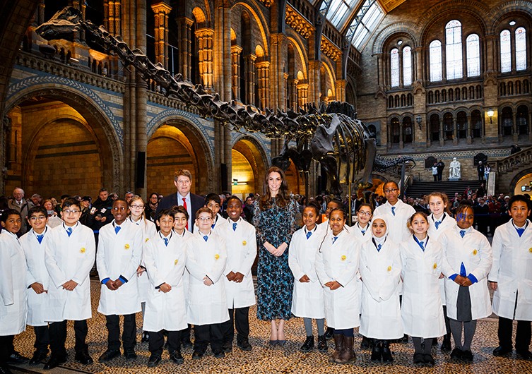 A group of children in white lab coats stand with a woman in a blue dress inside a grand hall, featuring a large dinosaur skeleton exhibit. The setting is bright and historical with high arched windows. The mood is educational and formal.