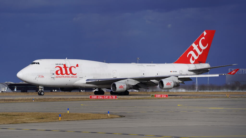 Boeing 747 on a runway, featuring red tail and logo. The sky is overcast, and the runway appears expansive and empty.